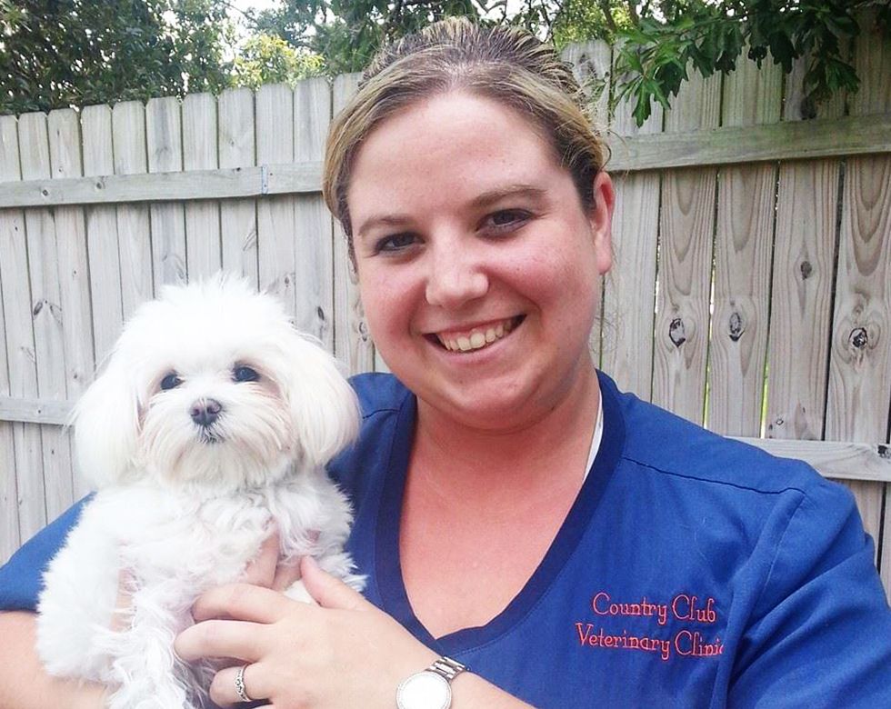 A smiling woman in veterinary scrubs holding a fluffy white dog.