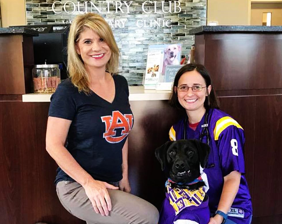 Two women in sports jerseys posing with a black dog in a clinic lobby.