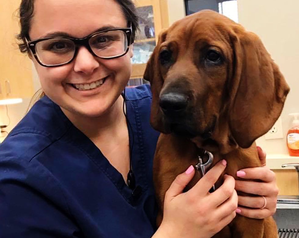 Smiling woman in blue scrubs holding a brown dog with floppy ears.